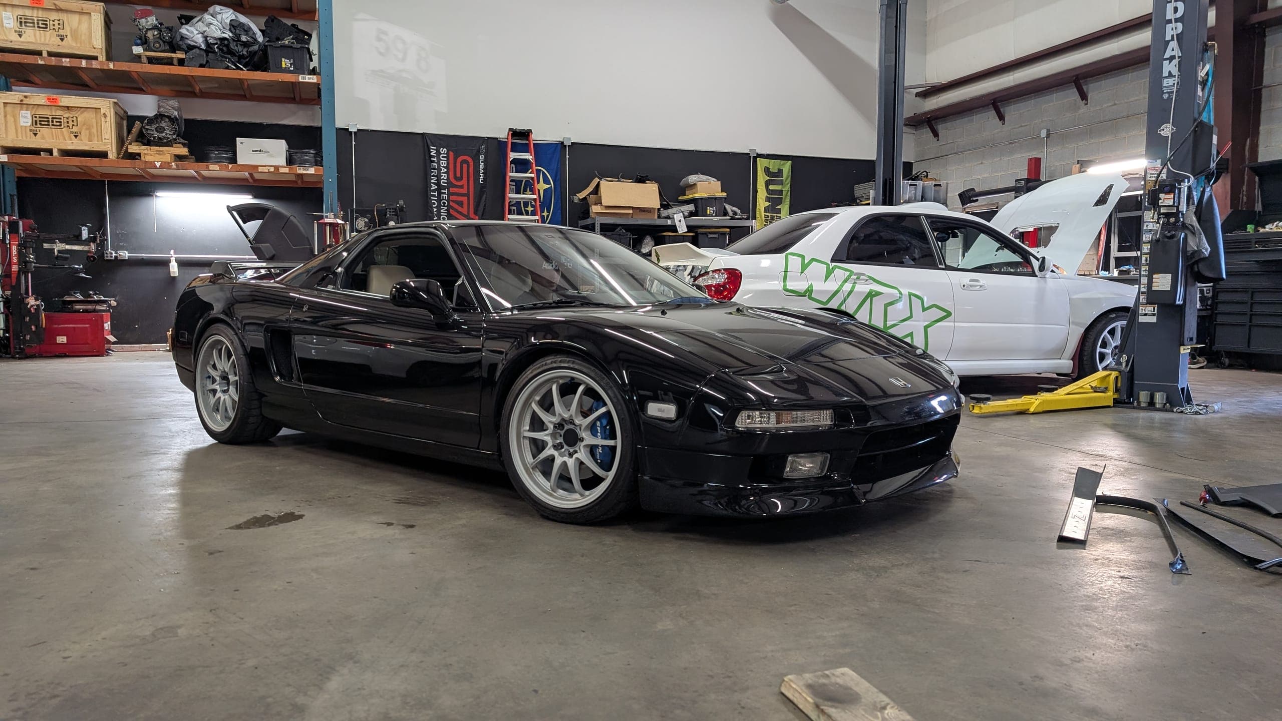 Black first-generation Acura NSX on the floor of the Streamline Performance Garage shop in Concord, NC, with a white Subaru WRX on the lift behind it
