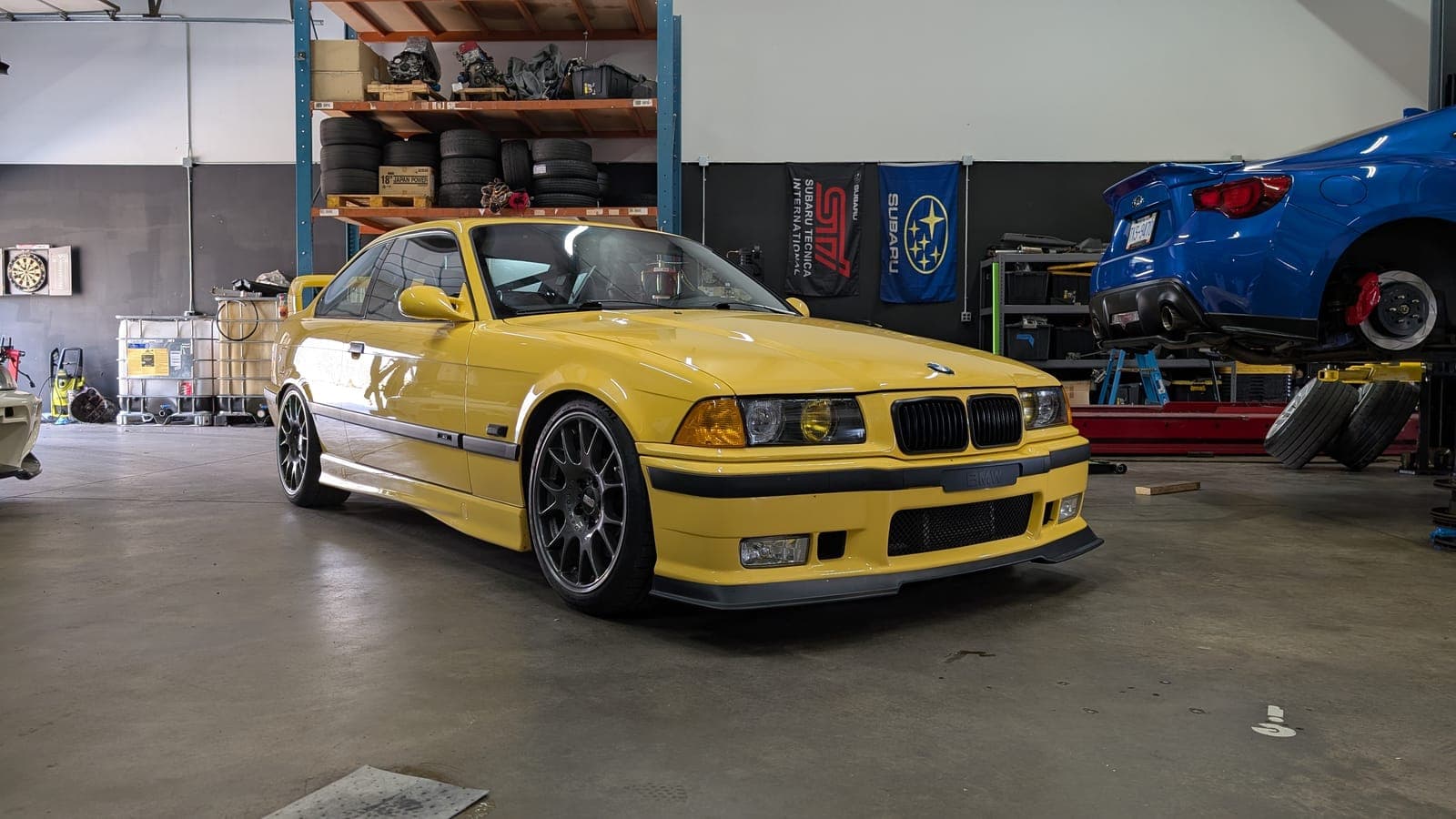 Dakar yellow BMW E36 M3 on the shop floor with a blue Subaru BRZ raised on the lift behind it