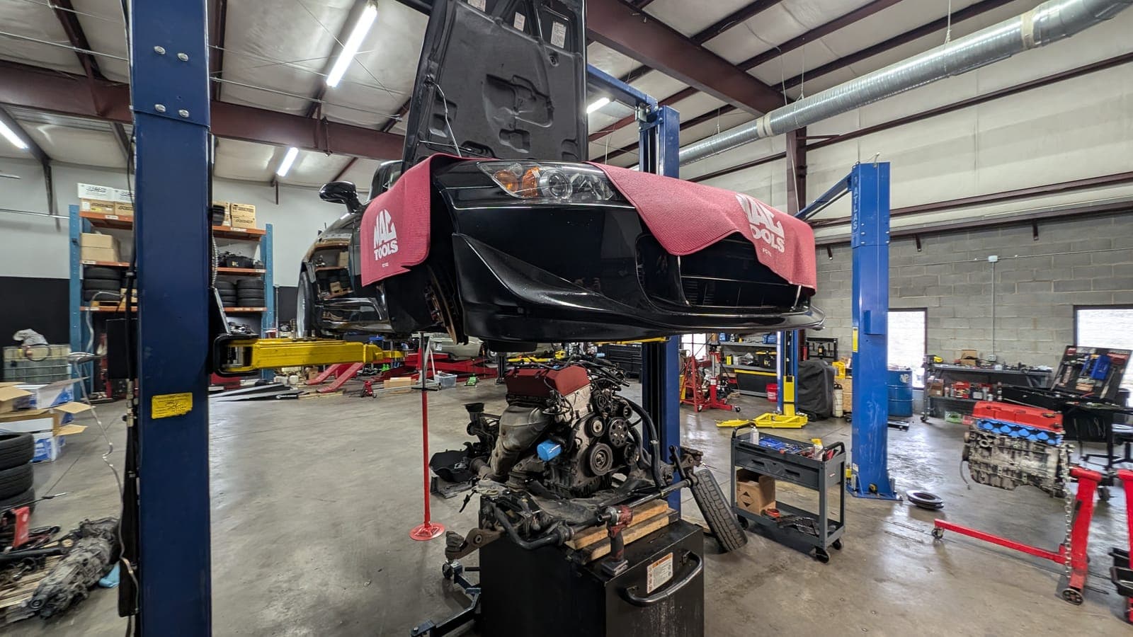 Car raised high on a two-post lift with its engine removed and sitting on a stand beneath it inside the shop