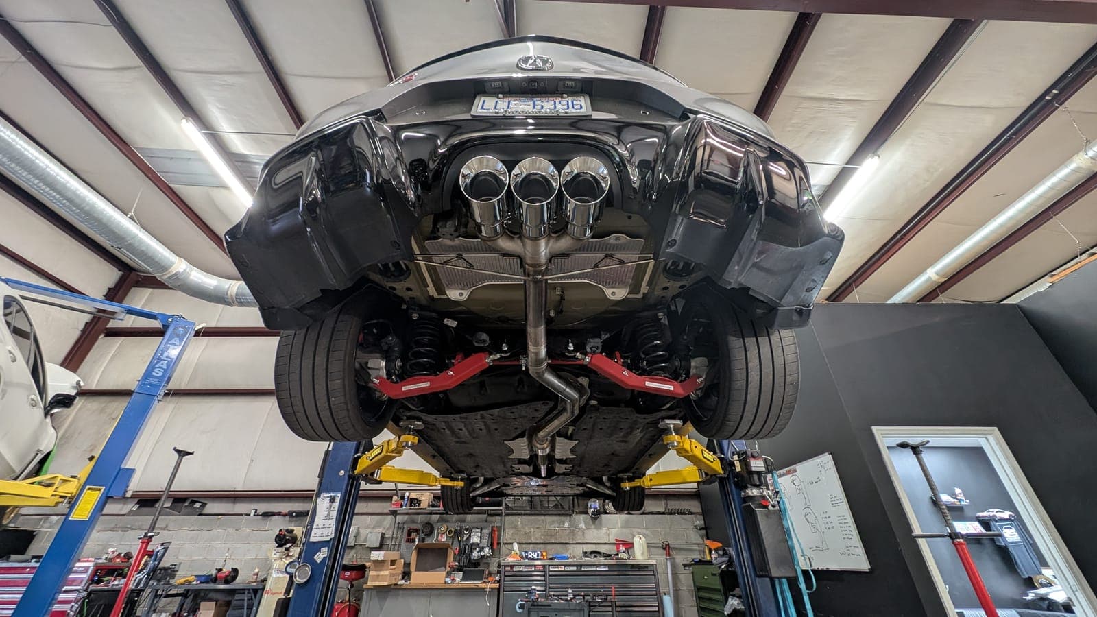 Underside of a black Lexus raised on the two-post lift, showing a center-exit exhaust with three tips and red rear suspension arms