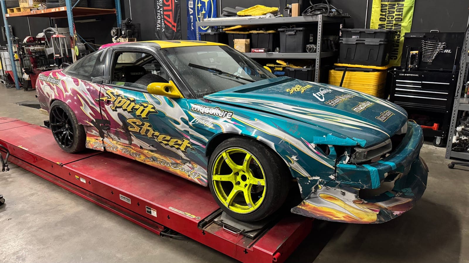 Nissan 240SX S13 drift car in a teal and pink Spirit Street livery on yellow wheels, sitting on the alignment rack with a cracked front bumper