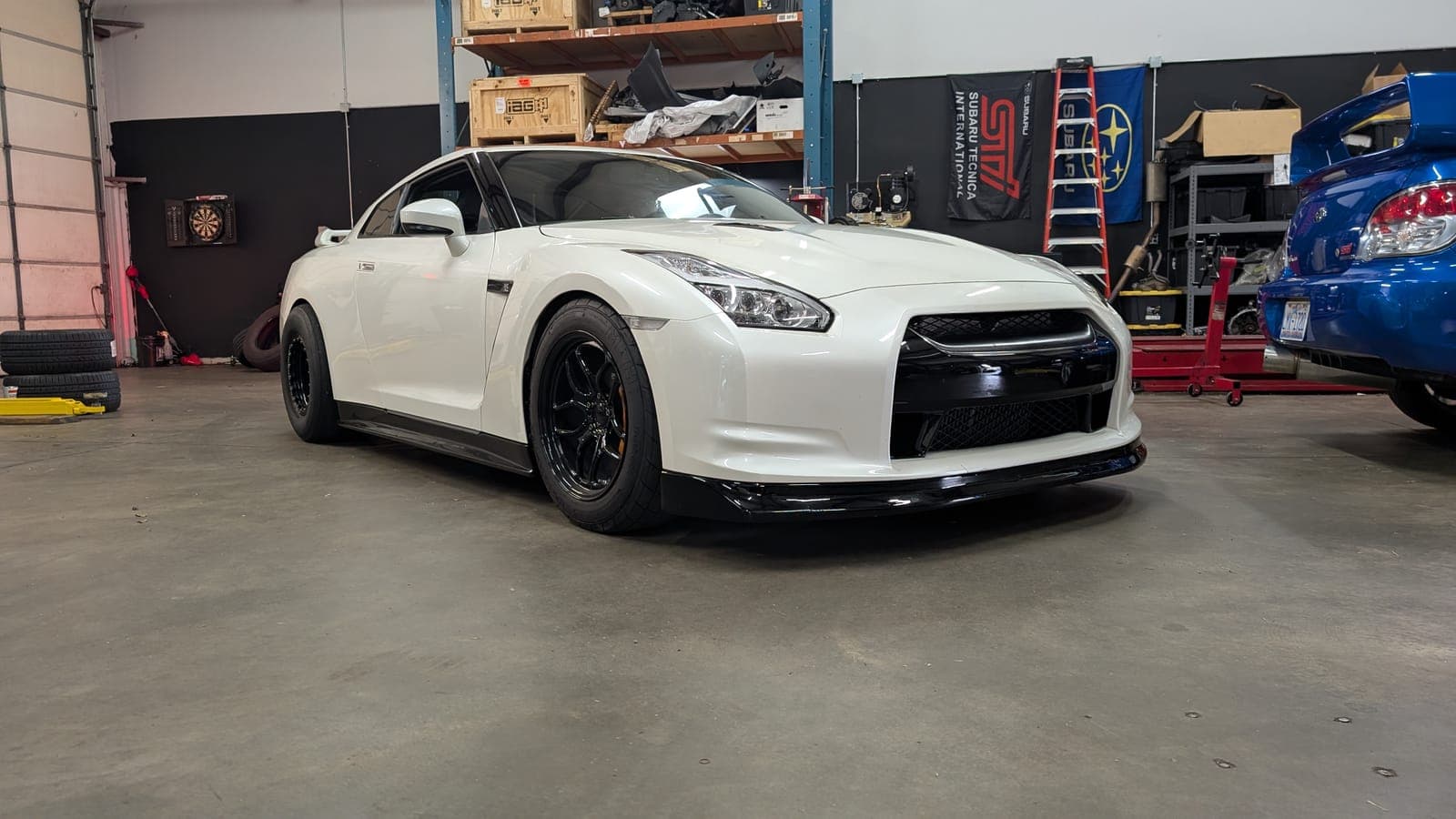 Pearl white Nissan GT-R R35 on black wheels on the shop floor with a blue Subaru WRX STI beside it