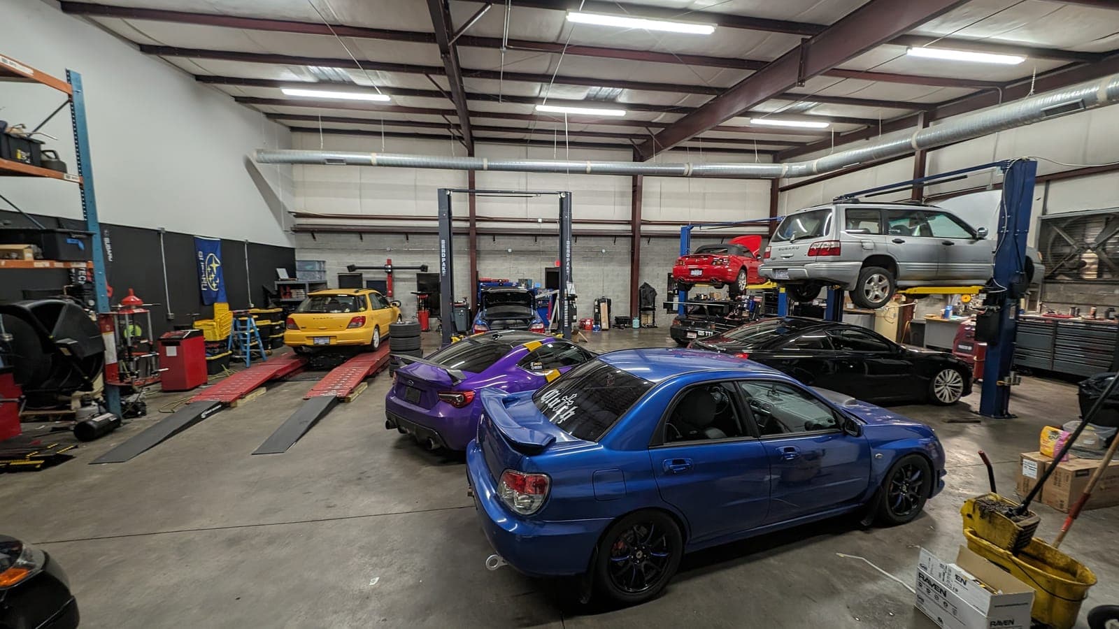 Busy shop floor with a blue bug-eye Subaru Impreza WRX, a purple Subaru BRZ, a yellow Impreza WRX and a silver Subaru Forester raised on a lift at Streamline Performance Garage in Concord, NC