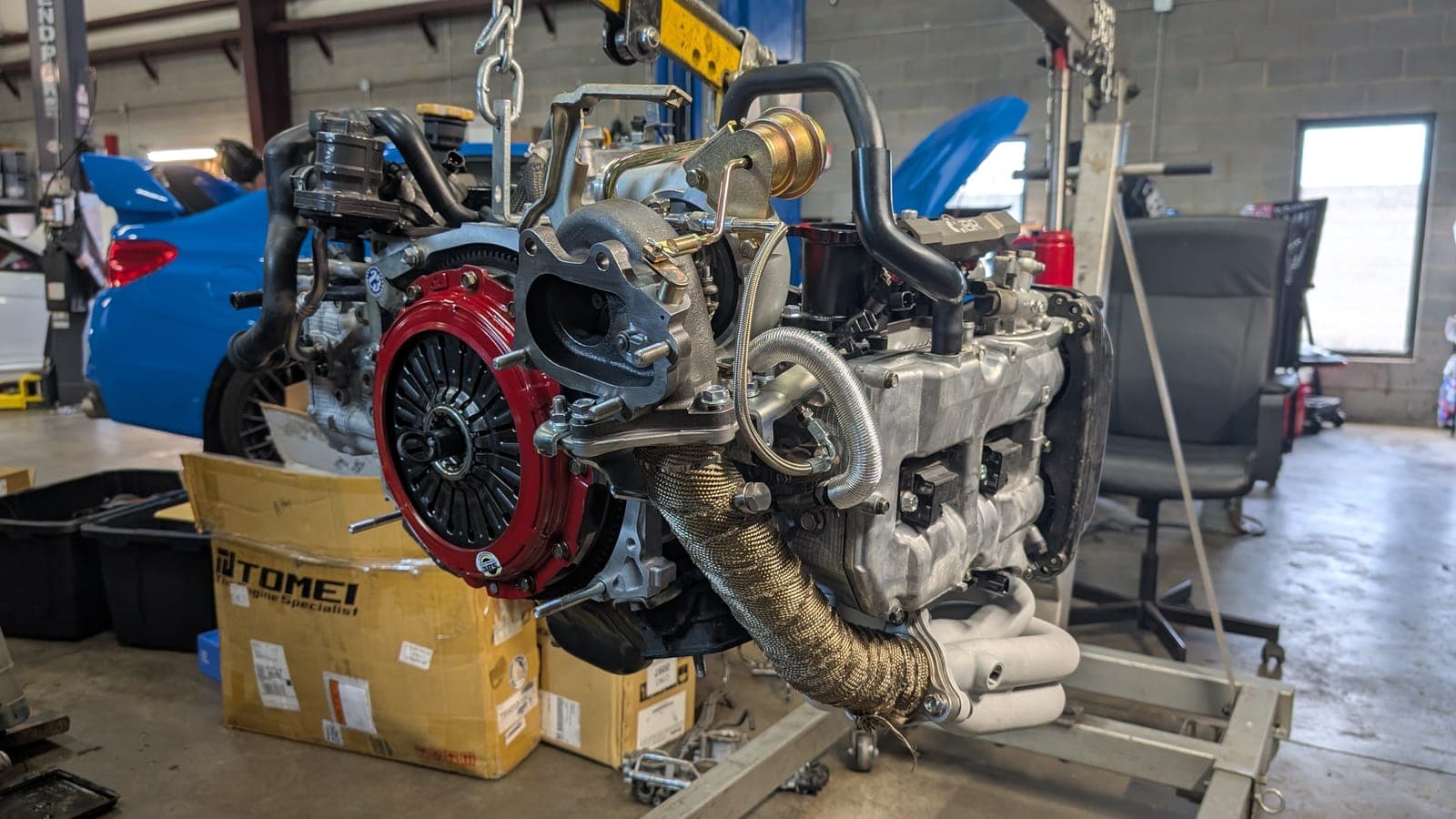 Built Subaru boxer engine on a stand with the turbo, heat-wrapped up-pipe, equal-length header and a red performance clutch fitted, with a blue Subaru WRX behind it