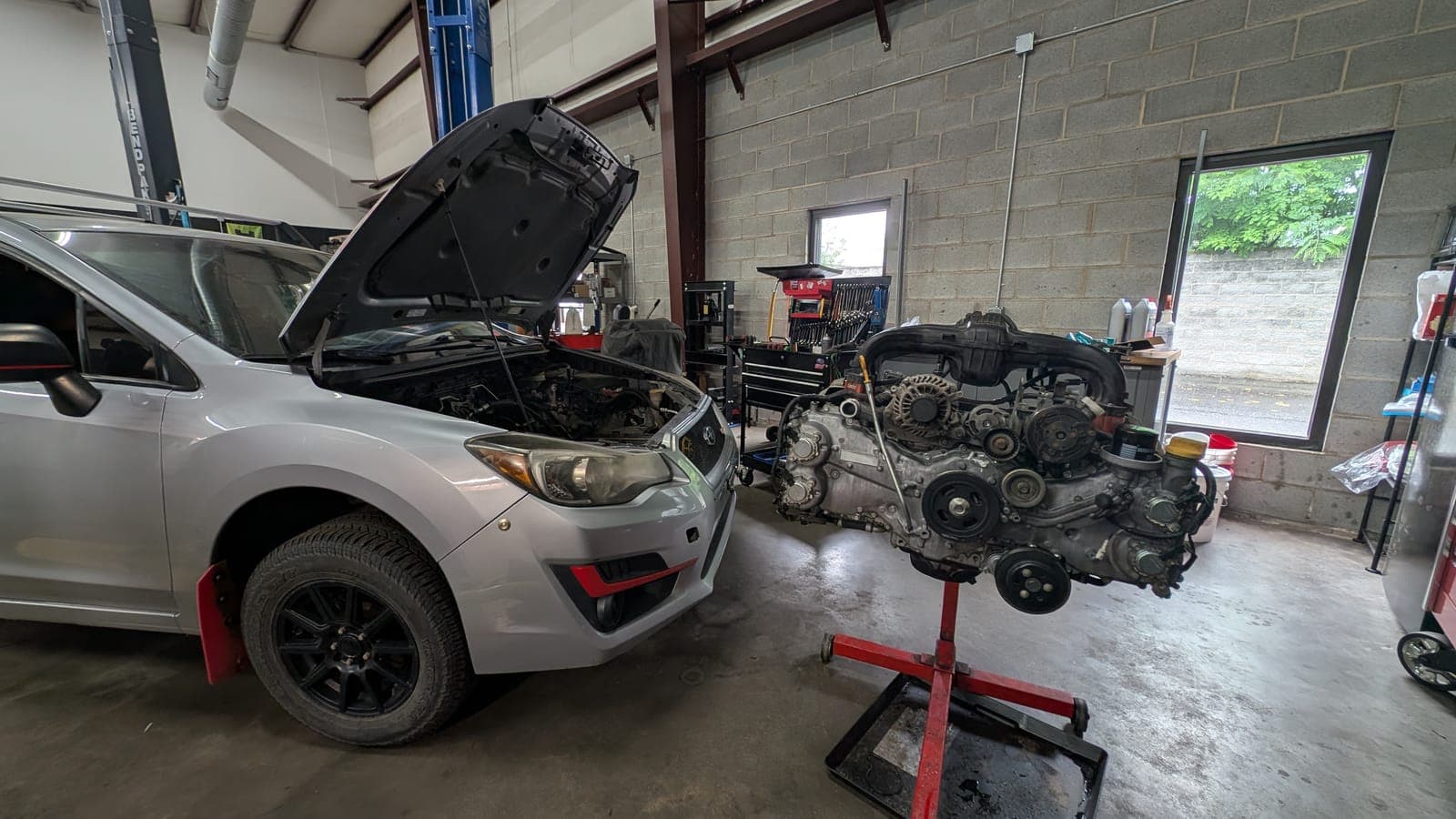 Silver Subaru Impreza with its hood open and its boxer engine on a stand beside it during an engine swap