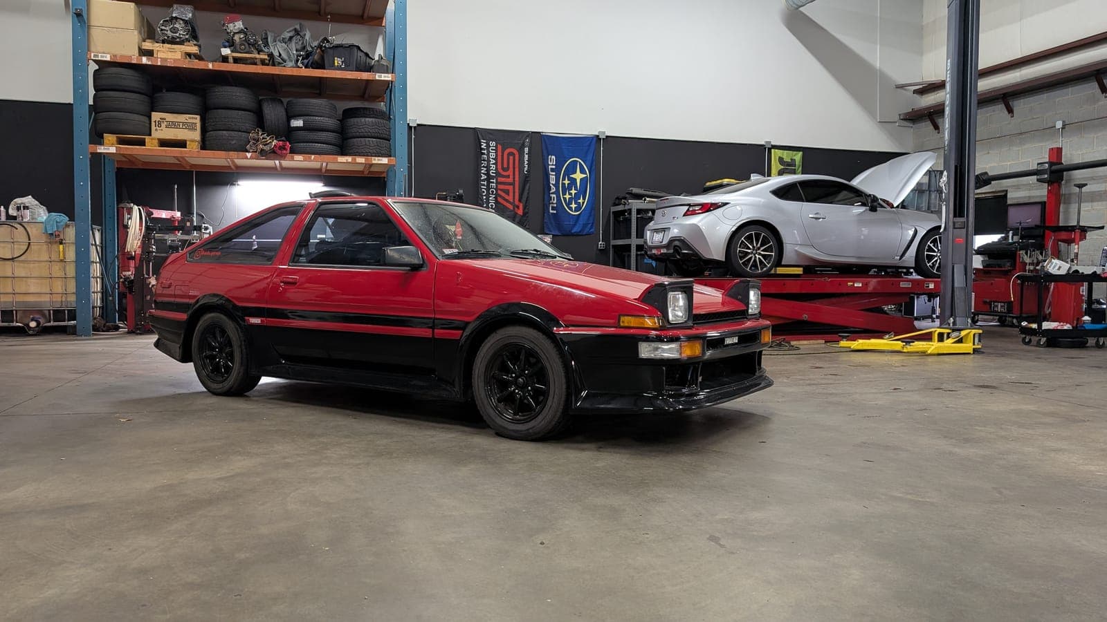 Red and black Toyota AE86 Corolla GT-S hatchback on the shop floor with a silver Toyota GR86 on the lift behind it, hood up