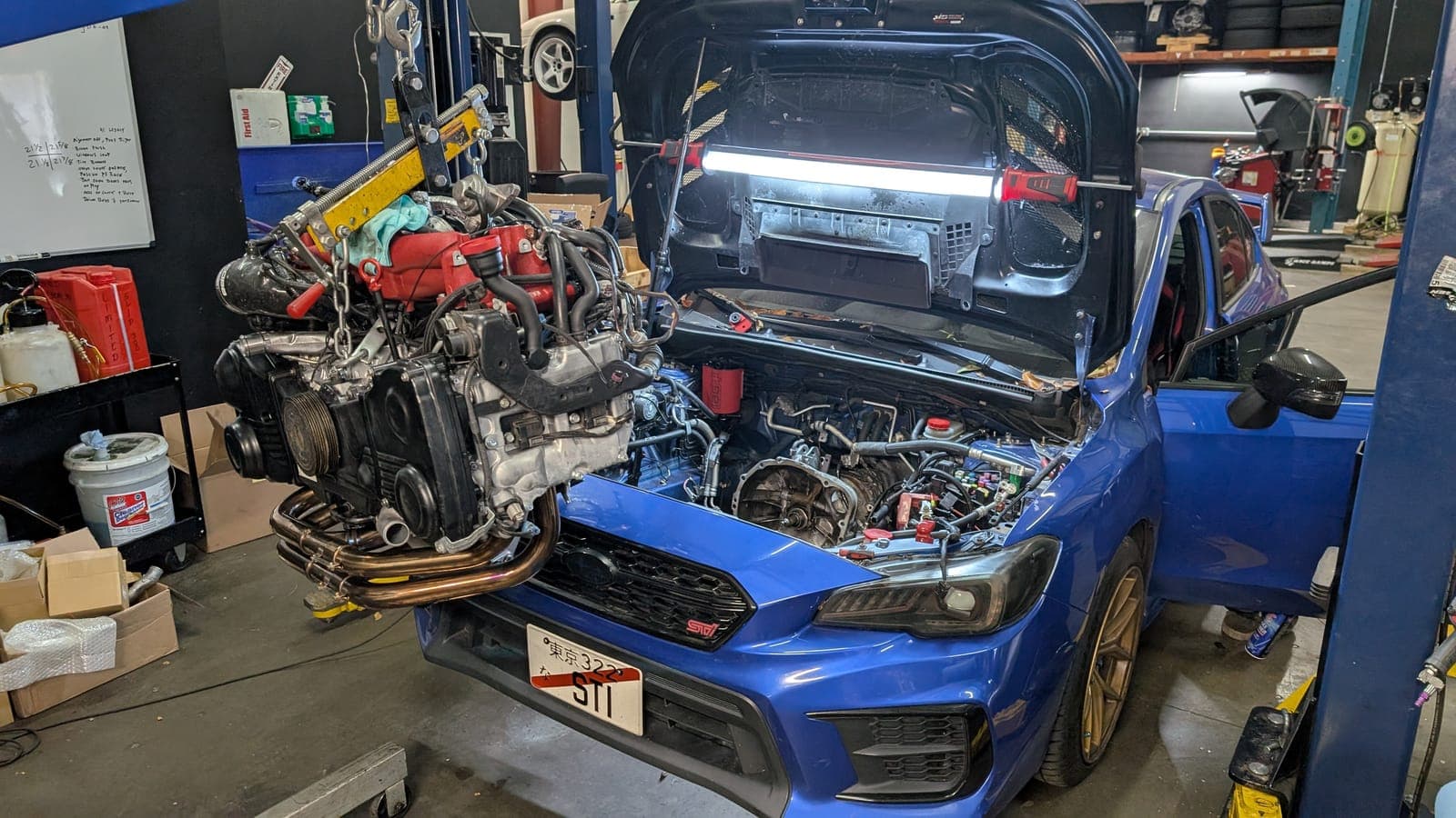 Blue Subaru WRX STI with its boxer engine removed and hanging from an engine hoist inside the Concord shop